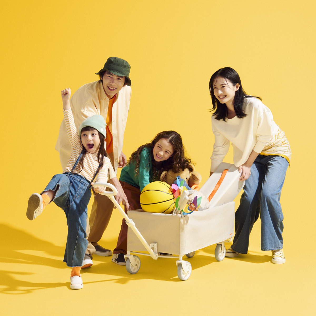 黄色い背景の中、おしゃれな白のキャリーワゴンを囲んで楽しむ4人家族。ボールやおもちゃを載せた、賑やかなアウトドアシーン