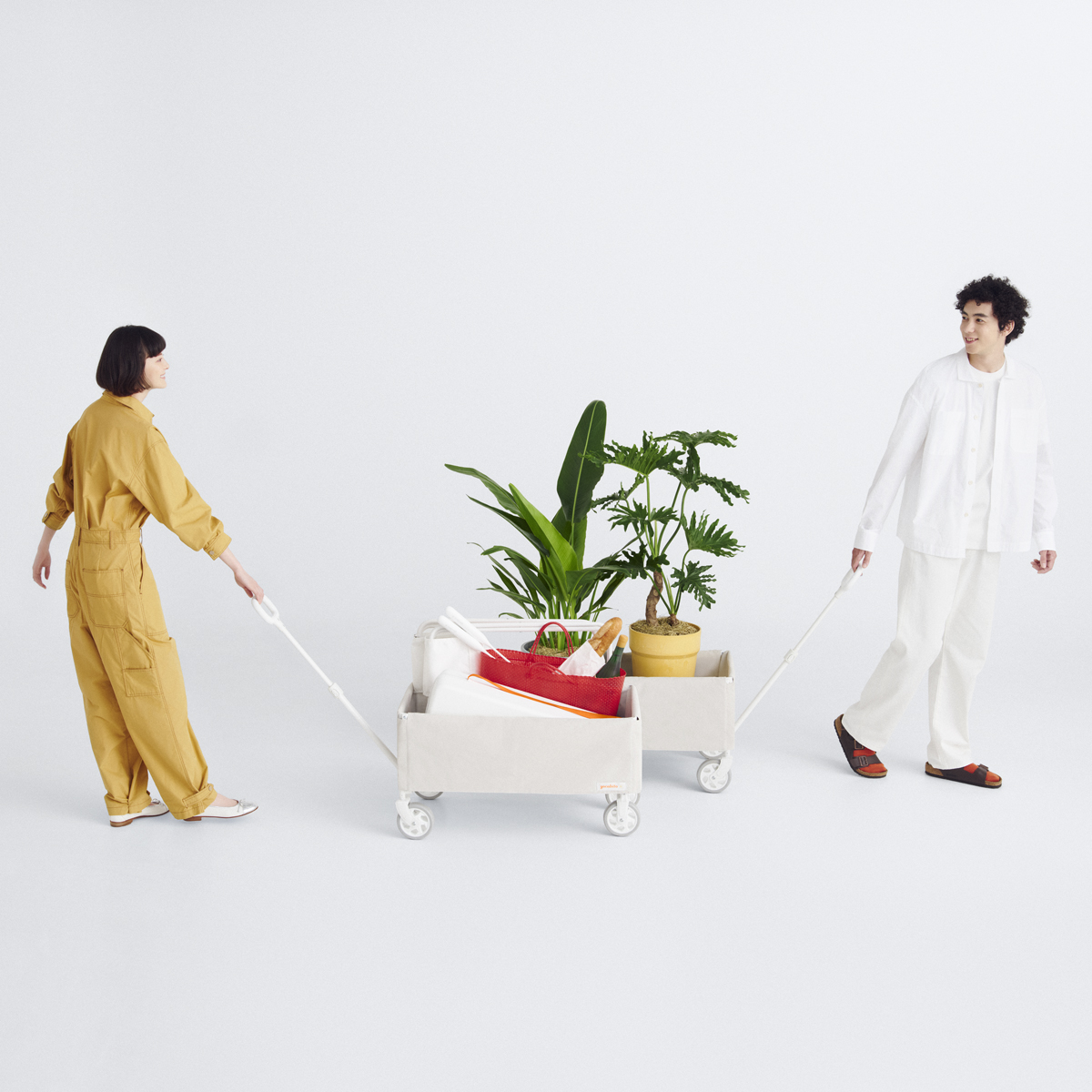 おしゃれな白のキャリーワゴンをそれぞれ引いてすれ違う男女。植物やピクニック用品を運ぶ、シンプルでスタイリッシュなアウトドアコーデ